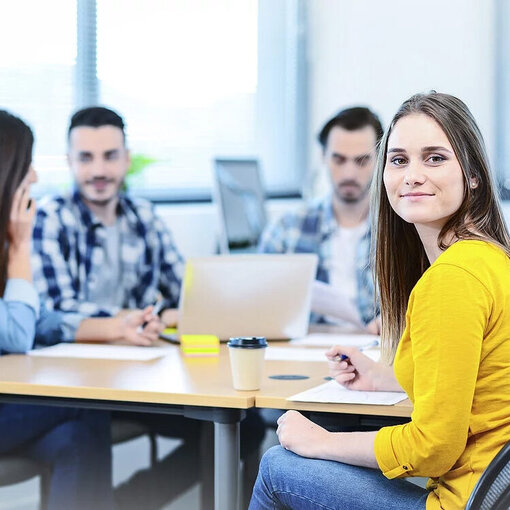 Junge Frau, die mit mehreren Personen an einem Tisch sitzt und in die Kamera lächelt
