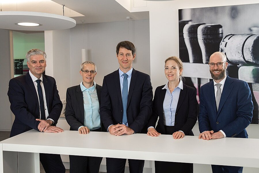 Unser Juristenteam Unser Juristenteam steht gemeinsam an einem Tresen und blickt freundlich in die Kamera